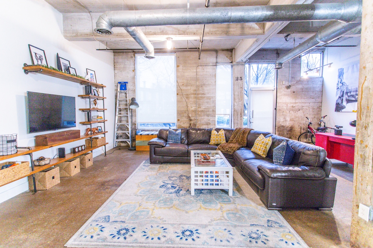 The spacious living area features a large sectional couch accompanied by textured throw pillows. A coffee table with a glass top sits on a decorative area rug. Wall-mounted shelves display framed photographs, and a 55" television is present, complemented by natural light from the sizeable windows.