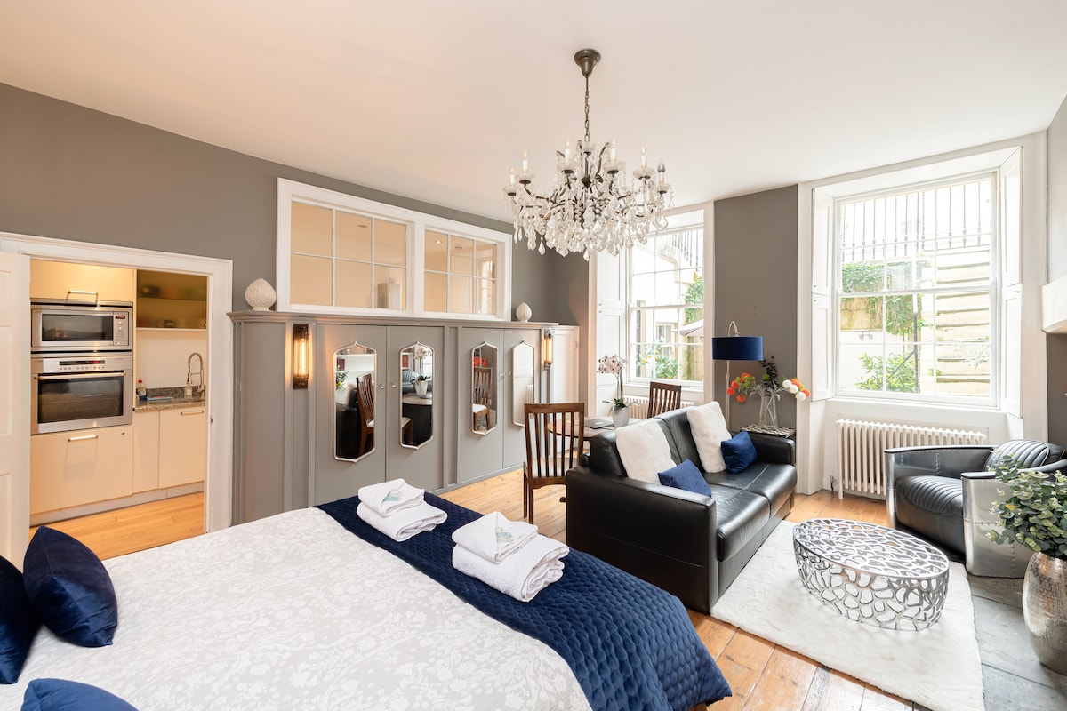 A spacious room features a large double bed with white linens and dark blue accents. A chandelier hangs above, illuminating the area. A leather sofa and a wooden dining table are present, with kitchen appliances visible in the background. Natural light streams through large windows.