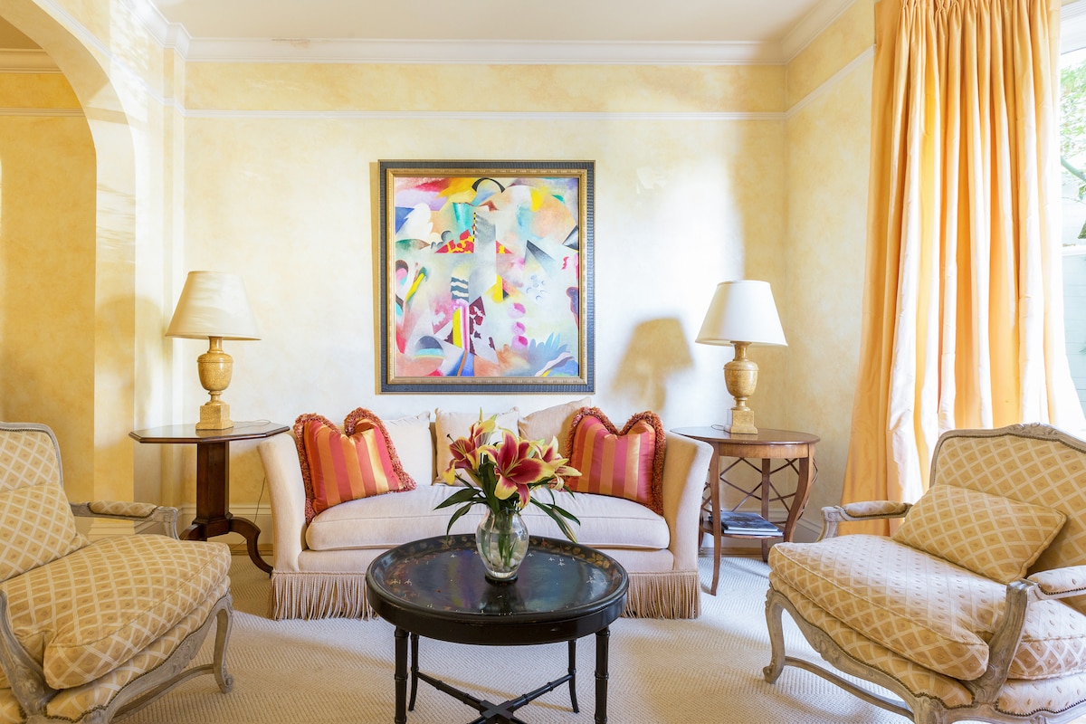 A cozy sitting area is presented with soft yellow walls, featuring a framed colorful artwork above a stylish sofa. Two patterned armchairs flank a round coffee table adorned with a floral arrangement, complemented by warm lighting from two table lamps.
