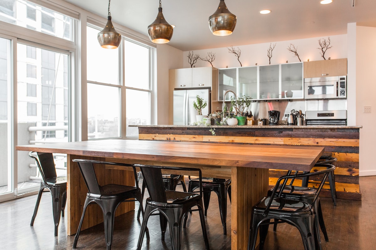 A spacious dining area features a long wooden table surrounded by black chairs. Modern pendant lights hang above, illuminating the room. A well-equipped kitchen is visible in the background, showcasing sleek cabinetry and various plants placed decoratively on the countertop.