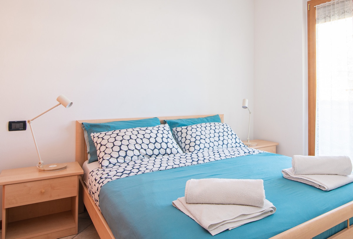 A spacious bedroom is furnished with a large bed featuring patterned bedding. Bedside tables are placed on either side, each with a lamp. Natural light enters through a window, with light curtains partially drawn.