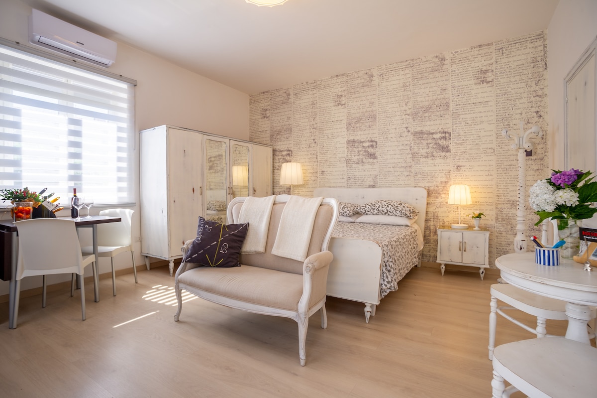 The bright studio features a cozy seating area with a light-colored settee. A spacious bed is framed by a wall decorated with text accents. A small dining table is positioned nearby, accompanied by two chairs. Natural light filters through a window with subtle blinds.