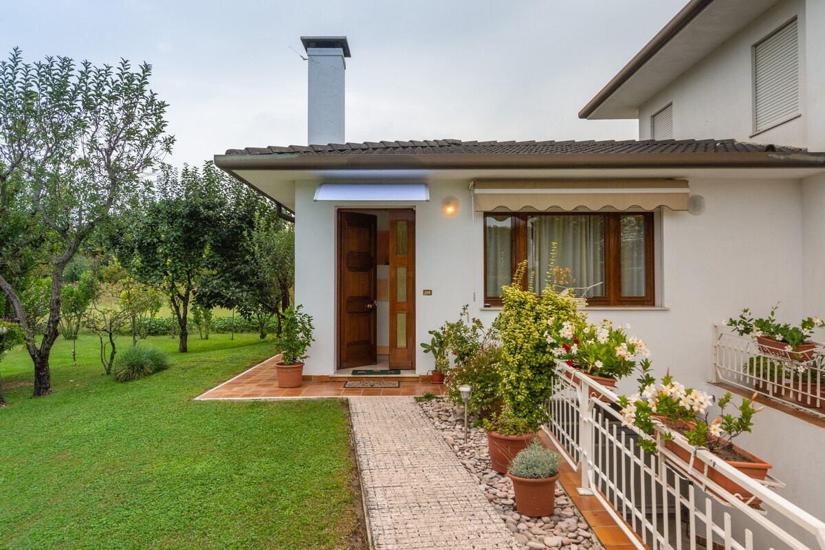 The entrance features a covered porch with a wooden door framed by plants in terracotta pots. The path, lined with pebbles, leads to an expansive green lawn with trees, offering a serene and welcoming environment.