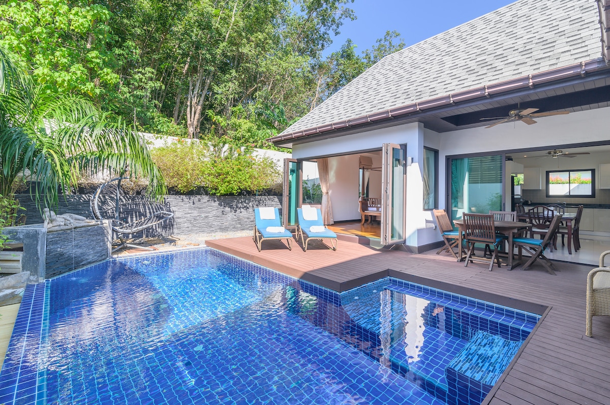 A modern villa displays a clear blue swimming pool, complemented by a jacuzzi area. Sun loungers are positioned beside the pool on a wooden deck. Lush greenery surrounds the outdoor space, enhancing the sense of privacy. Large doors open to an inviting interior area.