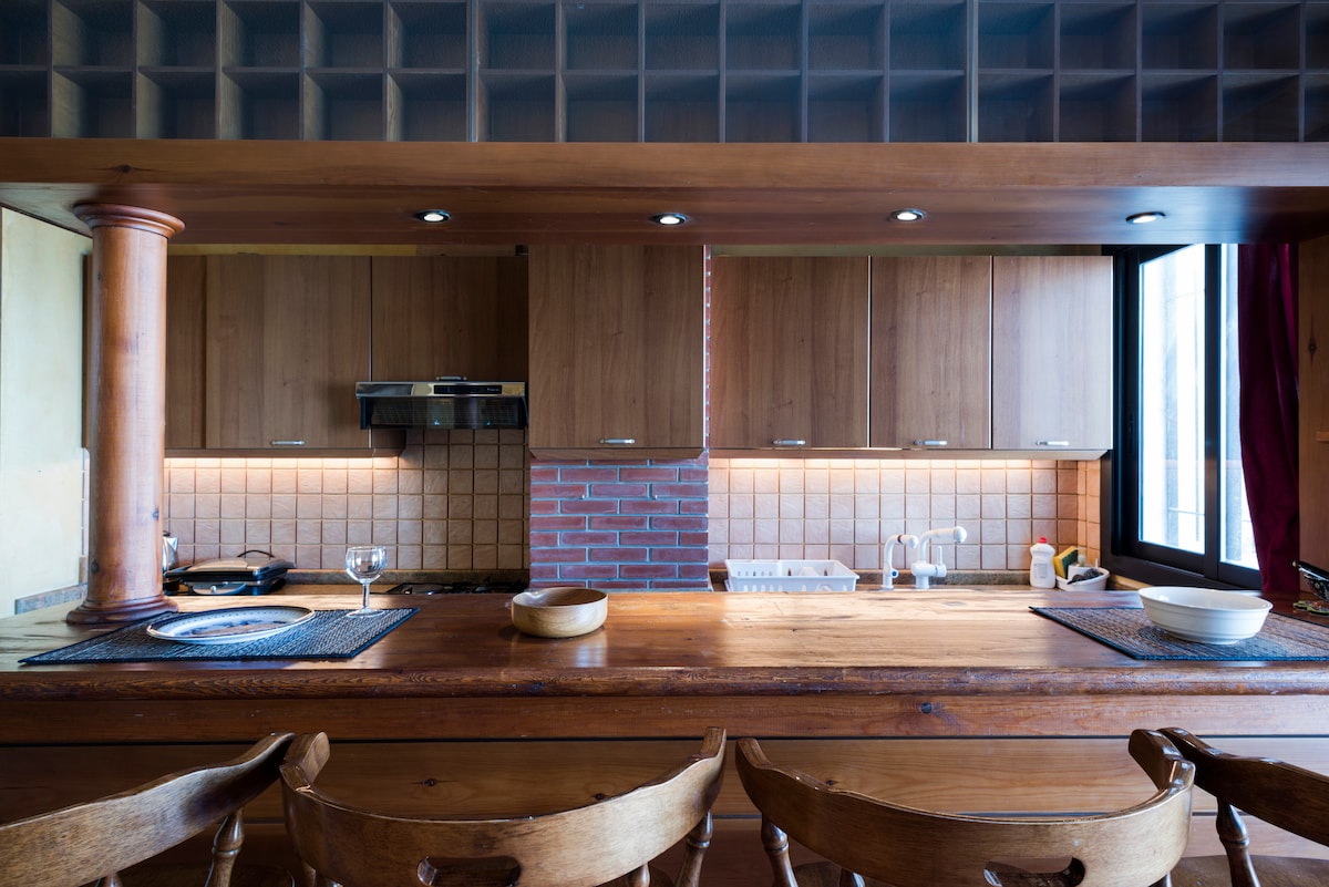 A modern kitchen is presented with wooden cabinetry and a combination of brick and tile backsplash. A spacious wooden countertop is accented by high-backed chairs, with warm lighting illuminating the area. A window allows natural light to enhance the surroundings.