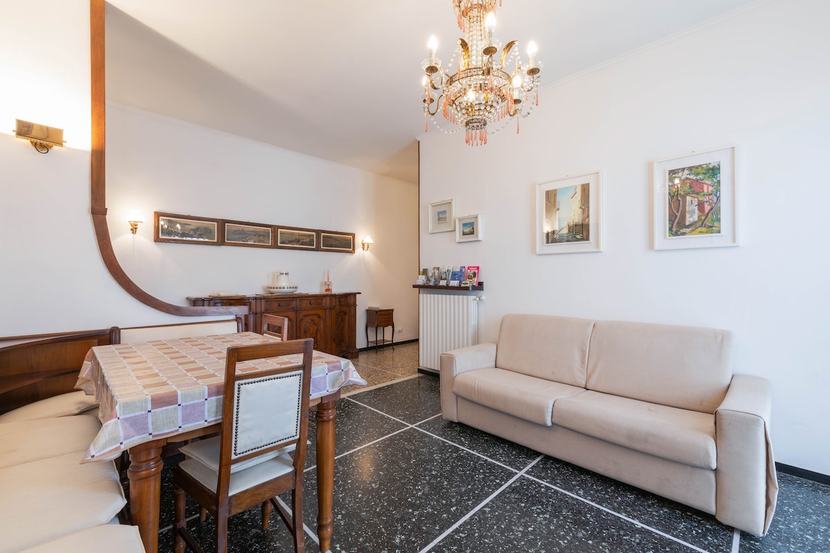 A spacious living area features a light-colored sofa and a dining table surrounded by wooden chairs. The floor showcases a classic black and white pattern. Artwork adorns the walls, and a chandelier hangs from the ceiling, providing ambient lighting.