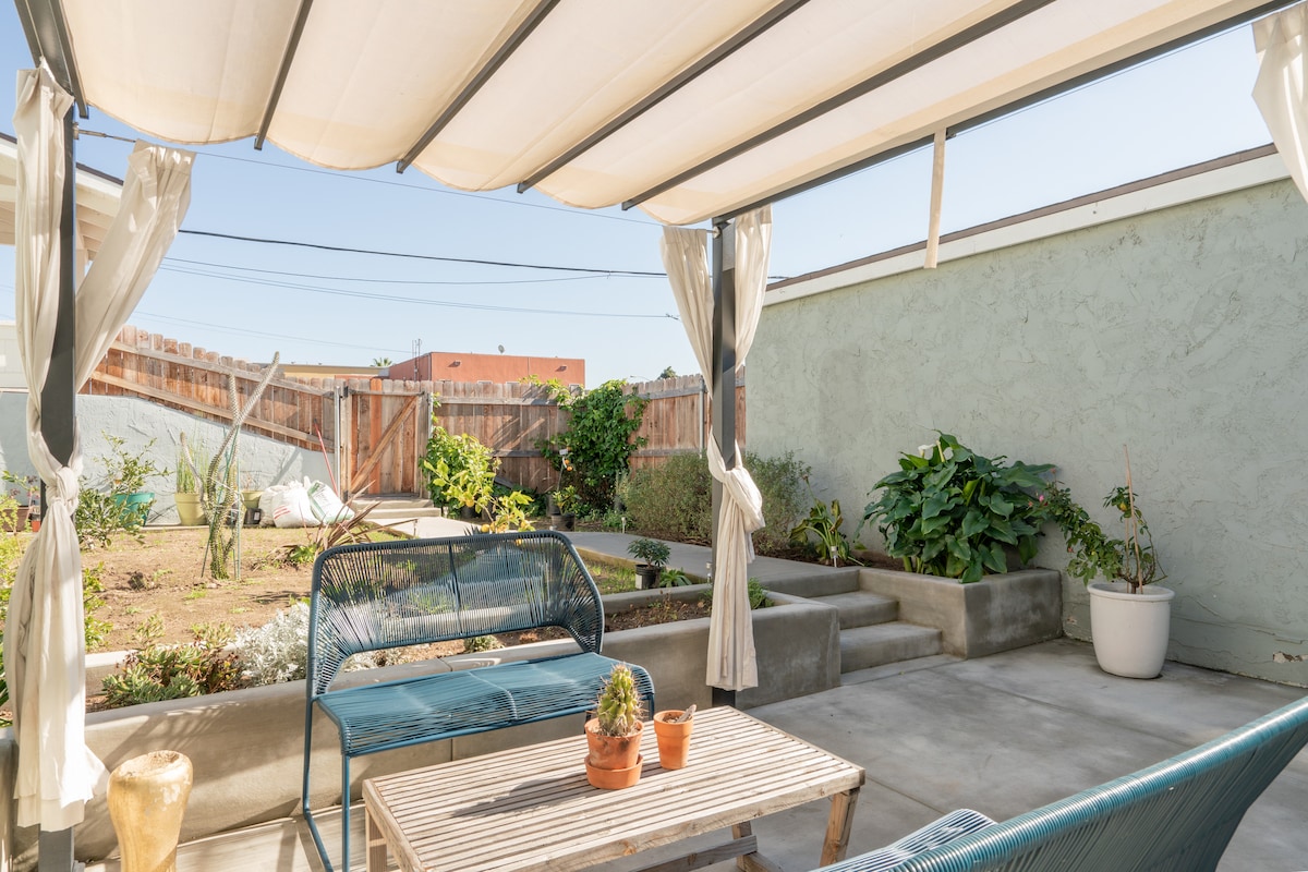An outdoor seating area is visible under a canopy, featuring a wooden table surrounded by two blue metal chairs. Potted plants are placed on the table, and a garden is seen in the background, bordered by a wooden fence.