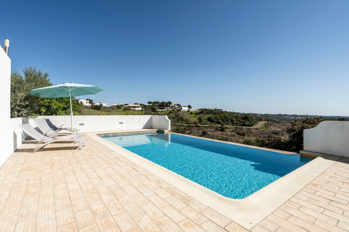 An infinity pool is framed by a sun terrace, featuring a light-colored stone surface. Two lounge chairs are positioned nearby, accompanied by a large umbrella providing shade. Rolling hills and clear blue skies serve as a backdrop, enhancing the tranquil outdoor space.