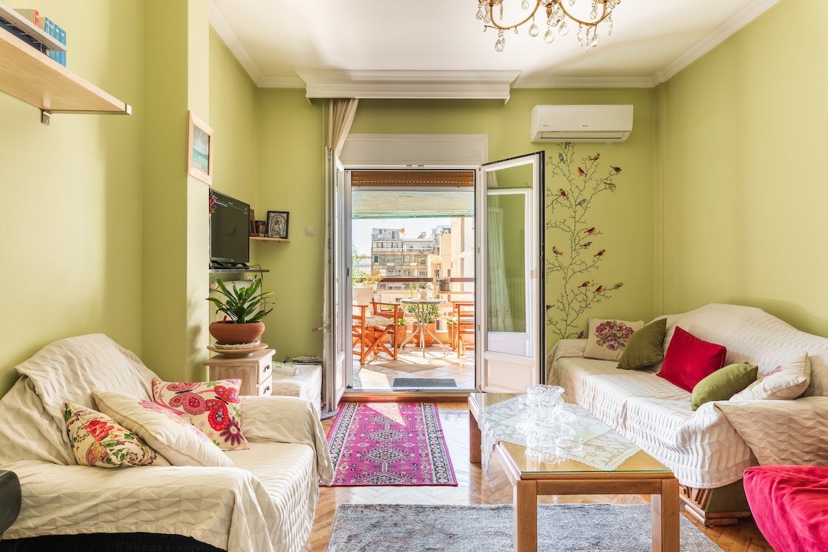 The living area features light green walls and two soft sofas with decorative pillows. A wooden coffee table is positioned in front of the sofas, with a colorful rug underneath. A bright balcony door opens to a terrace with outdoor seating, allowing natural light to fill the space.