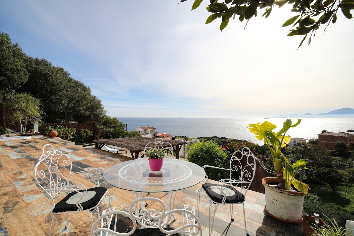 A scenic terrace features a glass table surrounded by white wrought iron chairs, showcasing views of the ocean and distant coastline. Lush greenery and potted plants enhance the outdoor space under a clear sky, inviting relaxation and enjoyment of the serene surroundings.