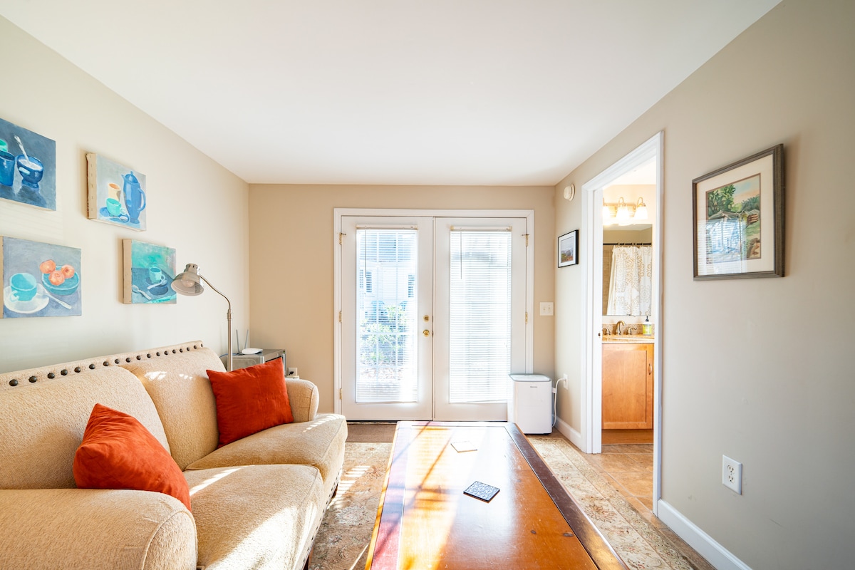 A brightly lit seating area features a comfortable couch with decorative pillows. A wooden coffee table occupies the center, complemented by artwork on the walls. Beyond sliding glass doors, a glimpse of outdoor space can be seen, with a doorway leading to the bathroom and kitchenette.