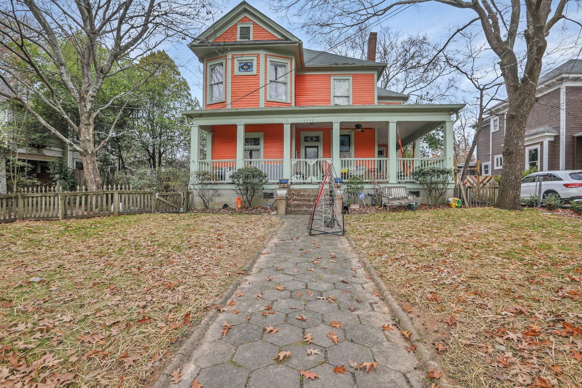 A charming Victorian house with a vibrant orange exterior is set against a backdrop of mature trees. A paved pathway leads to a welcoming front porch surrounded by greenery. The yard features a grassy area with fallen leaves, adding to the seasonal appeal.