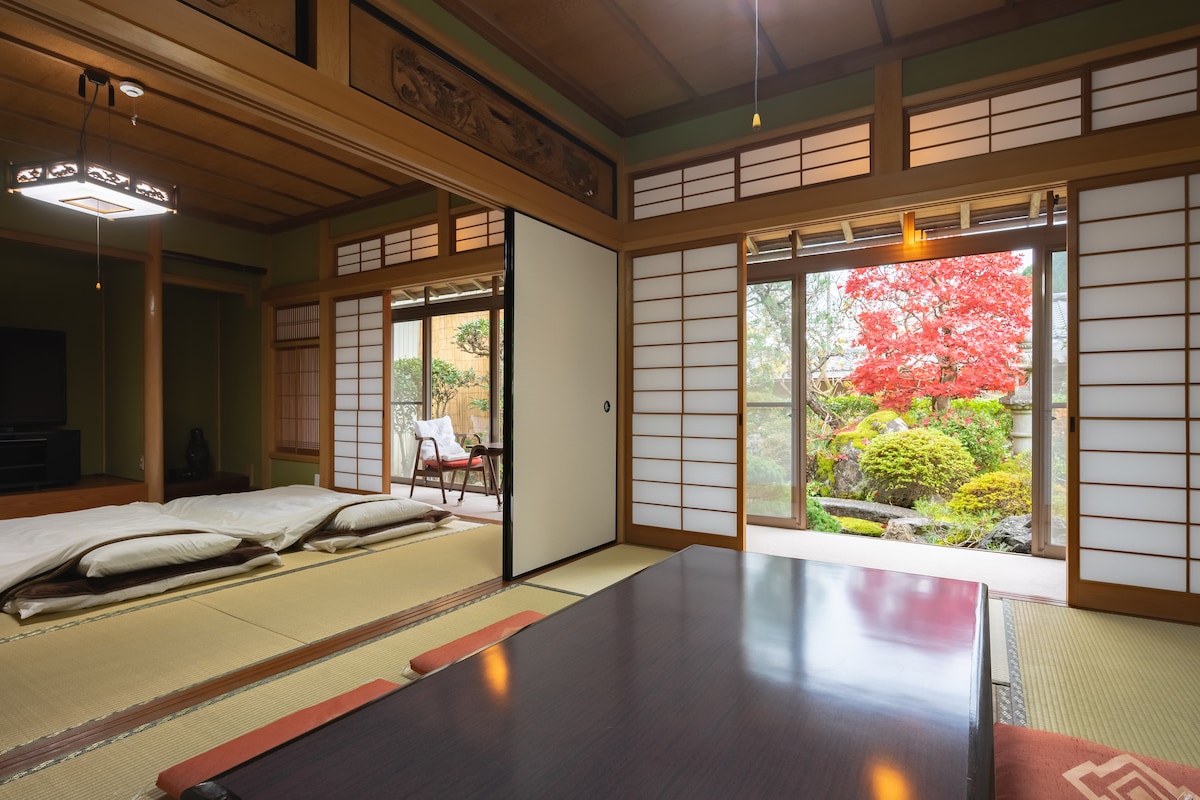 A spacious traditional Japanese room features tatami flooring and sliding shoji doors that open to a serene garden. Soft futons are neatly arranged on the floor, and a low table sits in the center. Natural light illuminates the space, highlighting vibrant colors from the outdoor foliage.