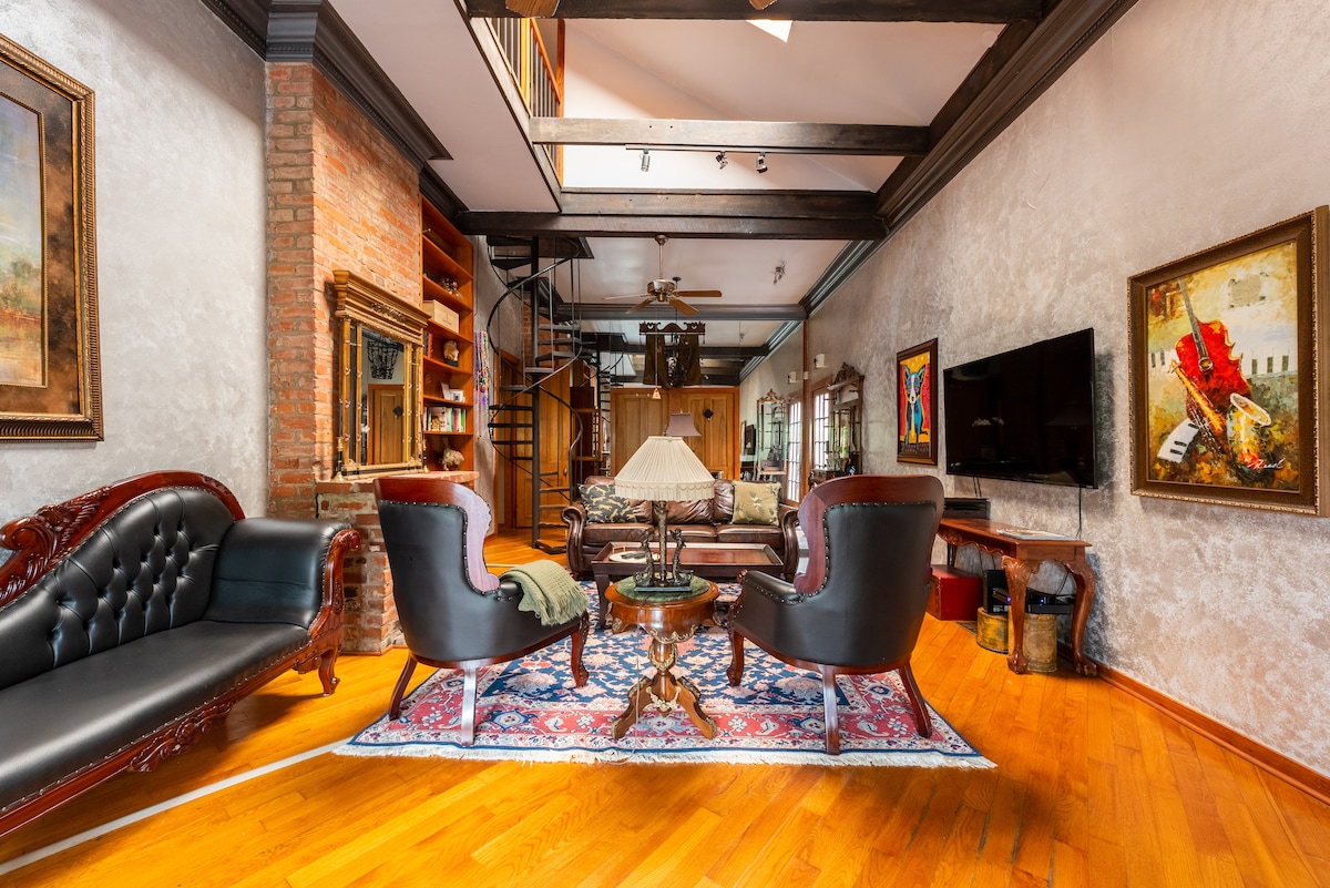 A spacious living area features a mix of classic and modern furnishings, including a dark leather sofa and two ornate chairs. A colorful rug anchors the space, while a large flat-screen TV is mounted on the wall. Natural light pours in through high windows, enhancing the warm wood floors.