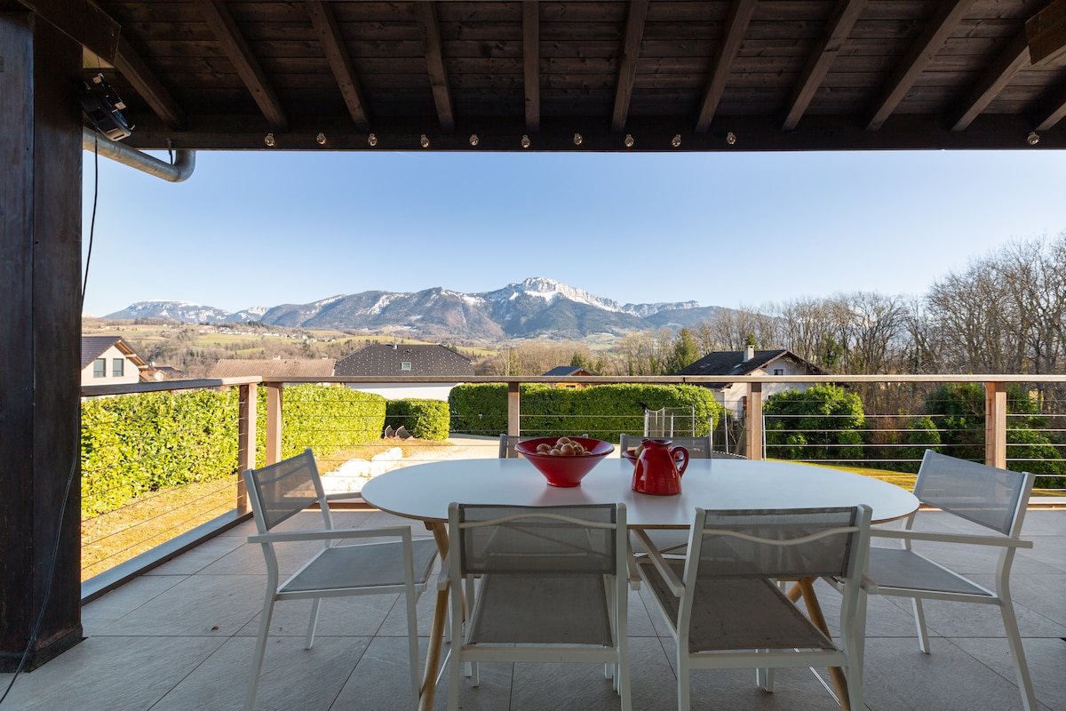 An outdoor terrace features a round table surrounded by several white chairs. A view of the snowy mountains is visible in the background, complemented by lush greenery. The setting invites relaxation and dining while enjoying the surrounding scenery.
