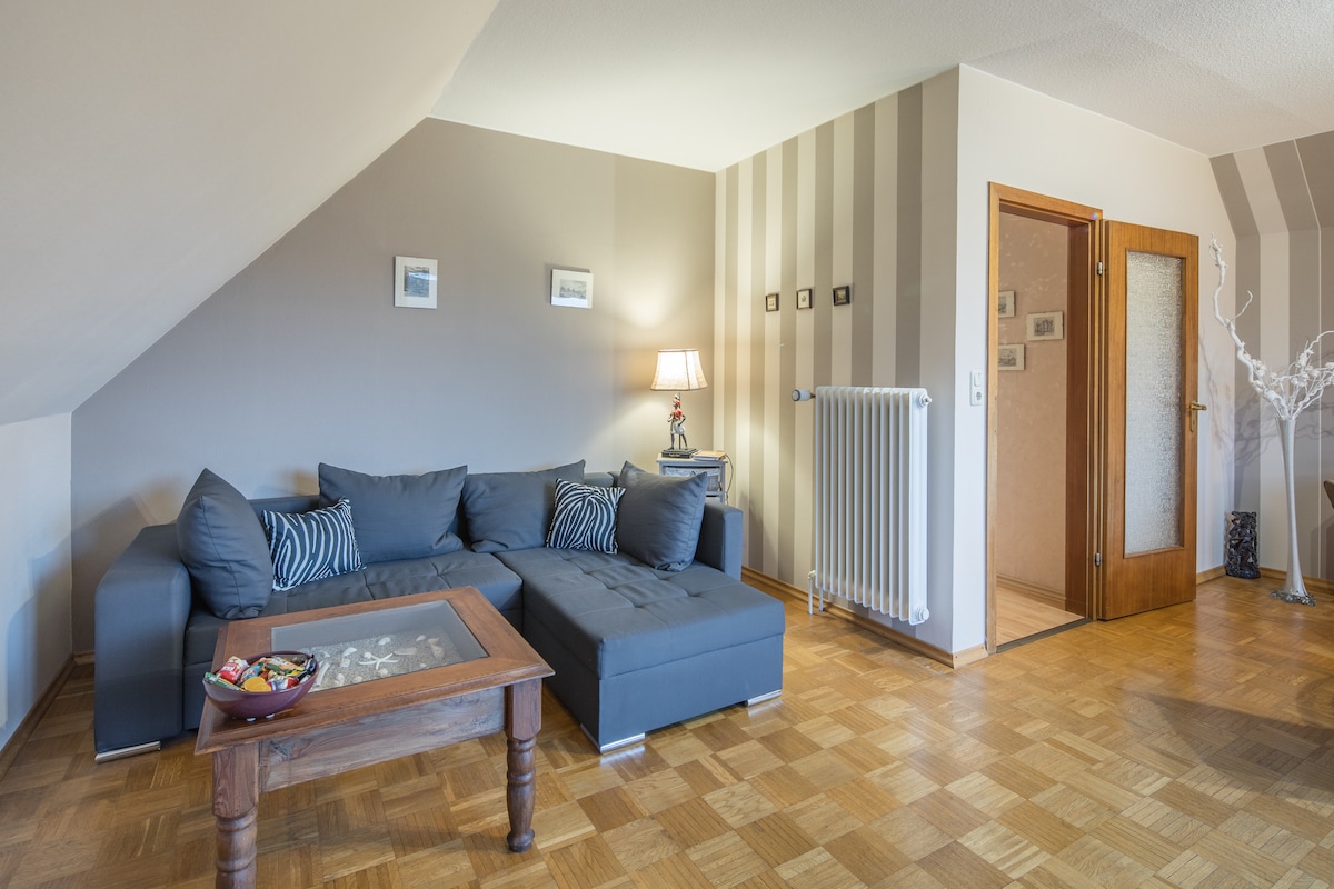 A modern living area features a comfortable sectional sofa with decorative cushions, positioned near a wooden coffee table. Soft lighting from a nearby lamp creates a welcoming atmosphere. The walls are adorned with framed pictures, and a doorway leads to additional rooms.