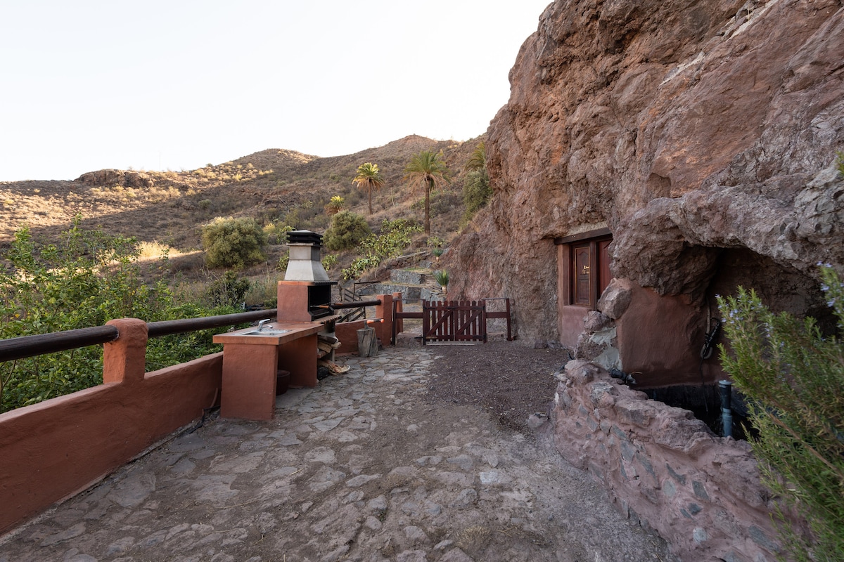 An outdoor patio area features a stone pathway leading to a barbecue grill, set against a rugged mountainous backdrop. A wooden fence encloses one side, and a natural rock formation provides an additional rustic element. Surrounding vegetation complements the serene environment.
