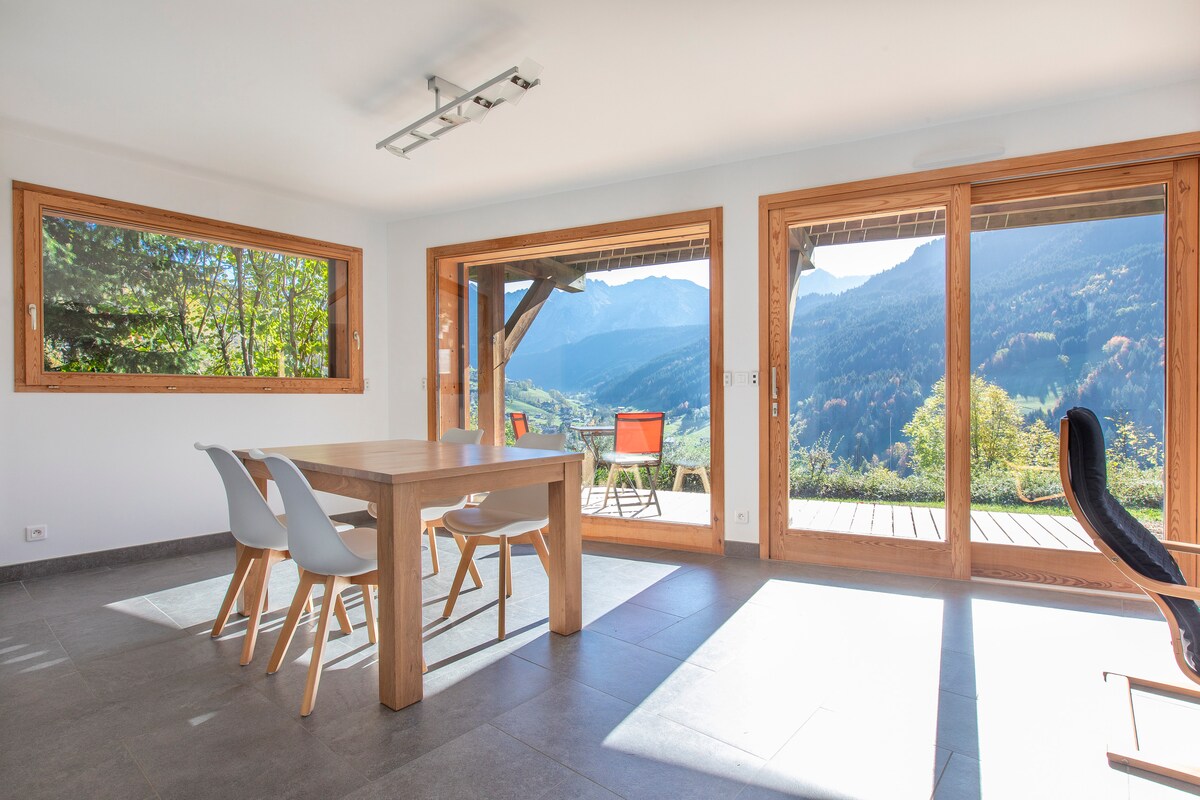 A bright dining area features a wooden table surrounded by six white chairs. Large windows provide expansive views of the mountains and outdoor terrace, allowing natural light to fill the space. The flooring is tiled, and the walls are painted in a neutral tone.