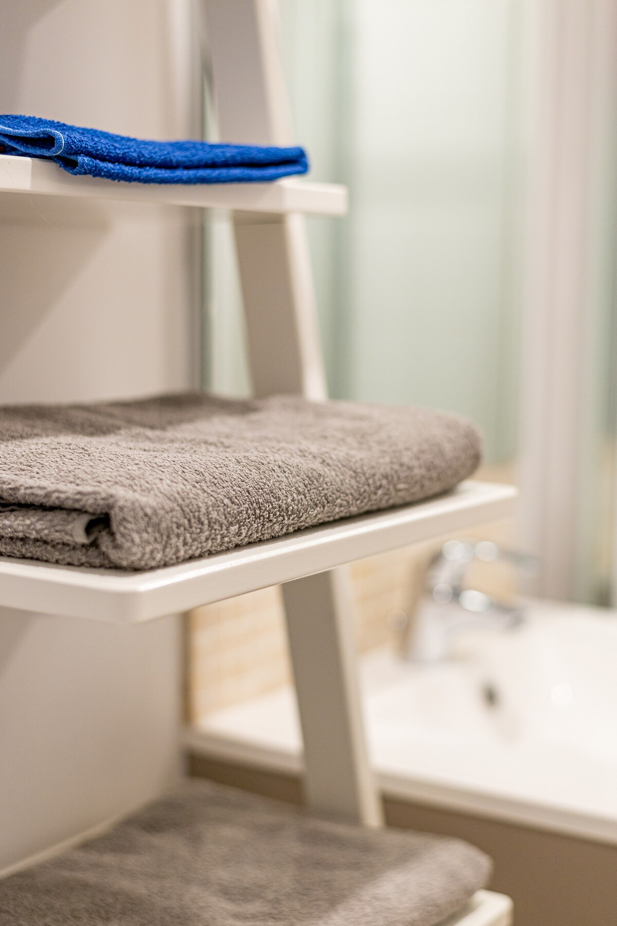 A neatly arranged bathroom shelf displays two folded gray towels, with one blue towel placed above them. The blurred background reveals a modern shower area and a glimpse of a washbasin.