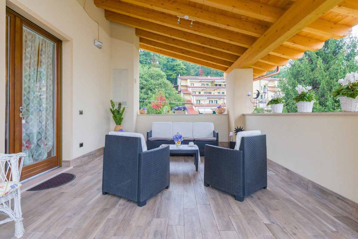An outdoor terrace is featured, showcasing a modern seating area with four chairs surrounding a low table. The space is shaded by a wooden roof, with green scenery visible in the background. Potted plants add a fresh touch to the space.