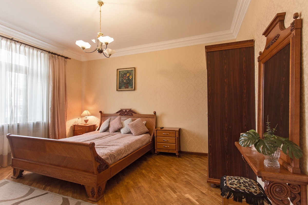 A spacious bedroom is presented, featuring a wooden bed with decorative pillows, matching wooden nightstands, and a tall wardrobe. Natural light enters through sheer curtains, illuminating the warm-toned walls and wooden flooring. A small plant is visible on the bedside table, enhancing the tranquil setting.