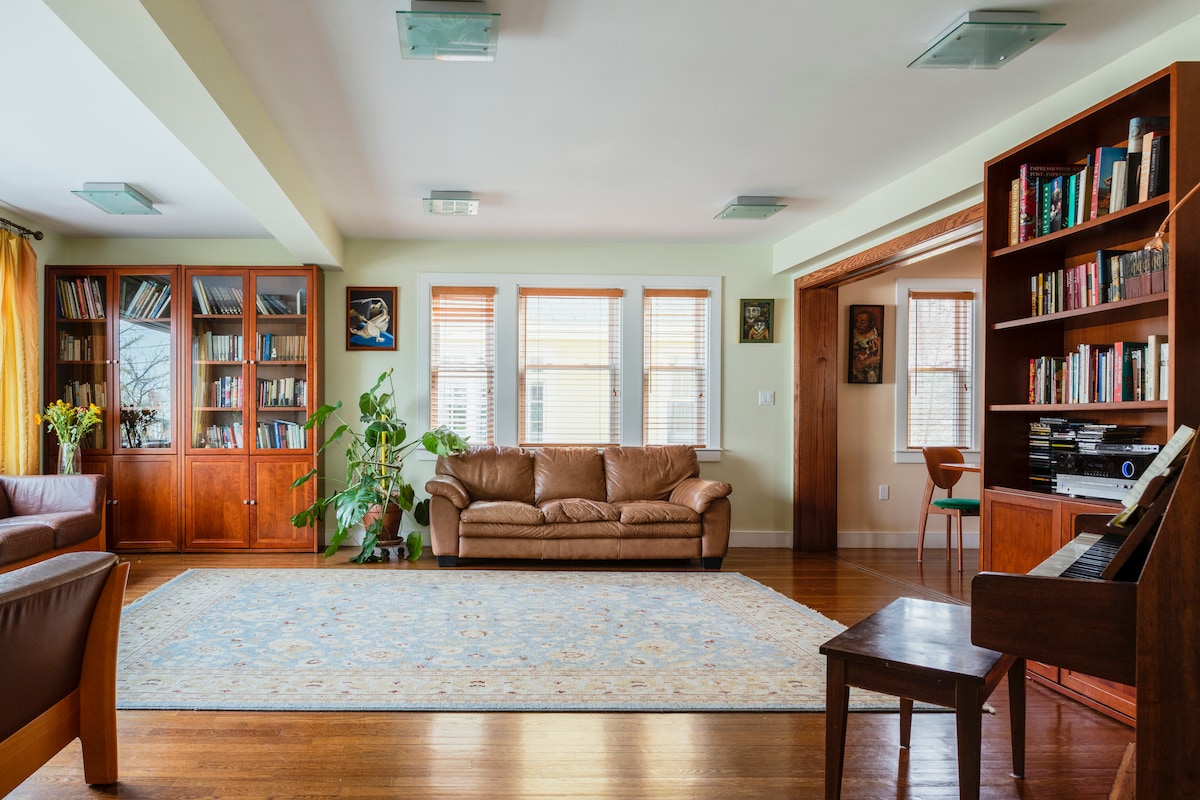 A spacious living room showcases a brown leather couch, surrounded by a bookshelf filled with books. Light filters through multiple windows, and a patterned rug covers the wooden floor. A small plant adds a touch of greenery, while a piano is positioned in the corner.