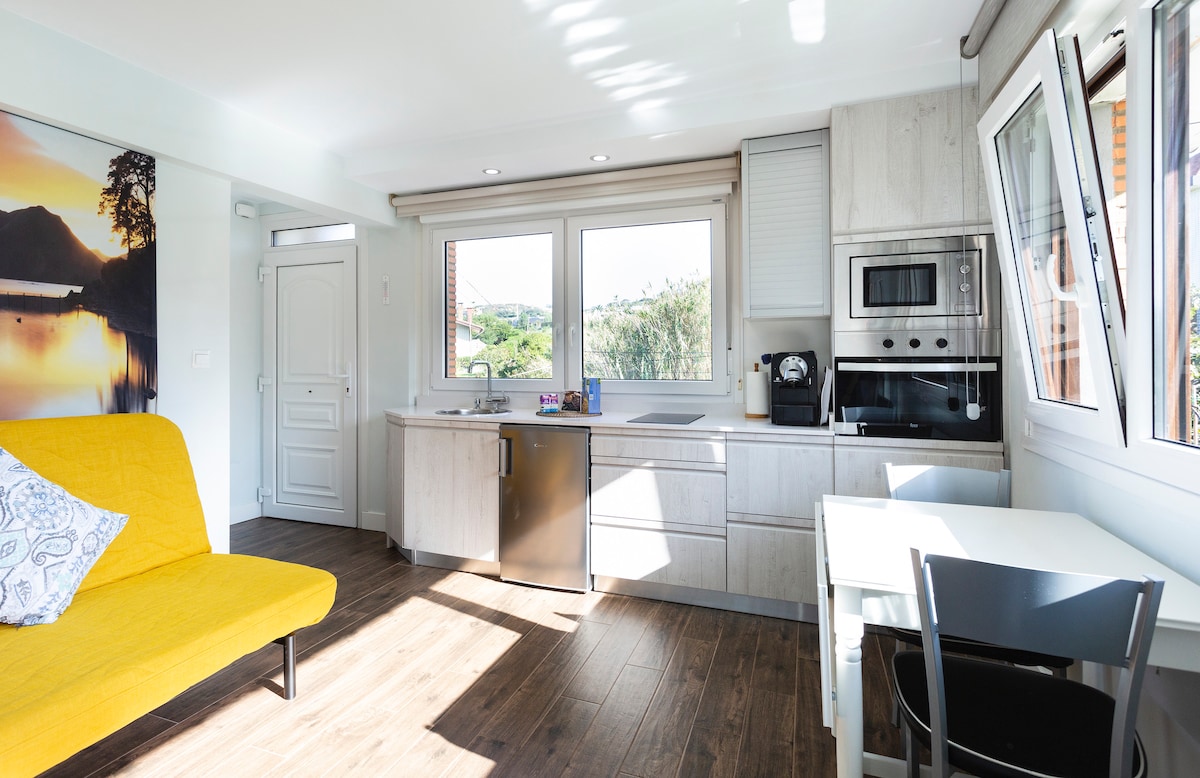 A bright kitchen area is equipped with modern appliances, including an oven, coffee maker, and refrigerator. Natural light filters through three large windows, providing views of the surrounding greenery. A small dining table and a yellow sofa bed complement the space, enhancing its functionality.