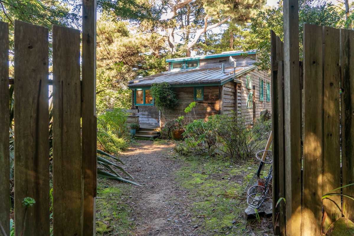 A welcoming entrance reveals a rustic house surrounded by lush greenery. Wooden gates open to a gravel path leading toward the home, flanked by native plants and trees. Sunlight filters through the foliage, enhancing the serene natural setting.
