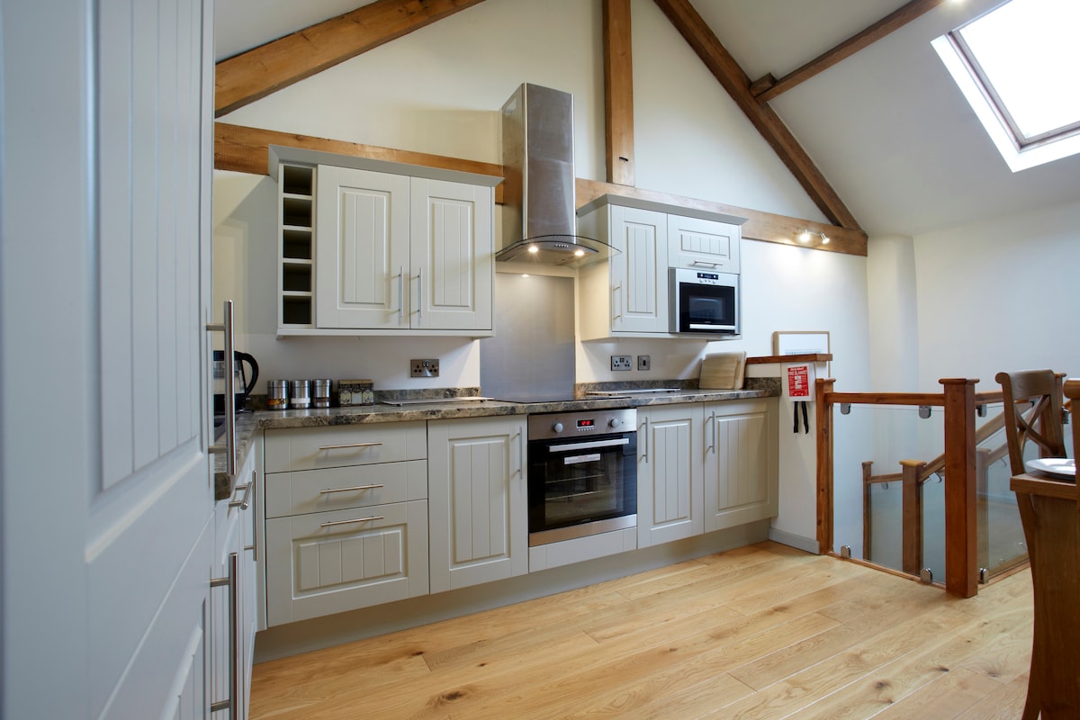 A modern kitchen is highlighted by light cabinetry and granite countertops, complemented by a stainless steel oven and hood. Natural light enters through a skylight above. Exposed wooden beams add rustic charm to the spacious design, enhancing the overall atmosphere.