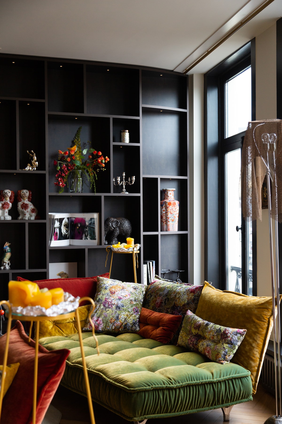 A vibrant seating area features a plush, multicolored sofa adorned with decorative pillows. A bookshelf filled with various decor items and books is positioned against the wall, providing an eclectic touch. Natural light pours in through large windows, illuminating the space.