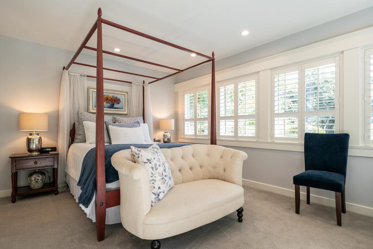A private guest suite features a four-poster bed with soft bedding, accompanied by a cozy tufted settee and a blue accent chair. Light enters through plantation-shuttered windows, highlighting the neutral color palette and creating a serene atmosphere.