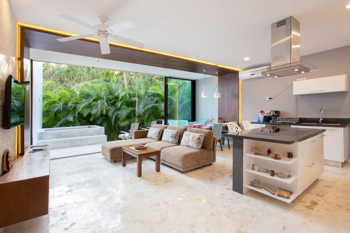 An open living area is highlighted by large windows that overlook lush greenery. A sectional sofa and coffee table sit comfortably in the space, complemented by a dining table nearby. A modern kitchen with sleek appliances and a breakfast bar is also visible.