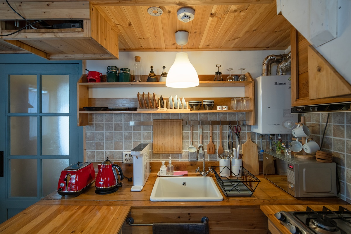 A warm wooden kitchen space is showcased, featuring natural wood cabinetry and countertops. Essential appliances, including a stove and a sink, are neatly arranged. Soft lighting is provided by a hanging pendant lamp, illuminating dishware and utensils on shelves. A cozy atmosphere is created by the rustic design.