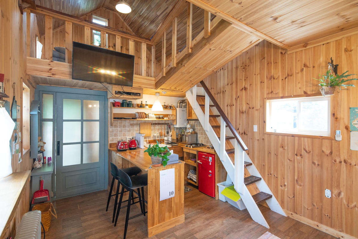 The interior of the wooden cabin features a warm wood paneling that creates a cozy atmosphere. A small kitchen area with essential amenities is visible, along with a wooden dining table and chairs. Stairs lead to a lofted sleeping area, and windows provide natural light.