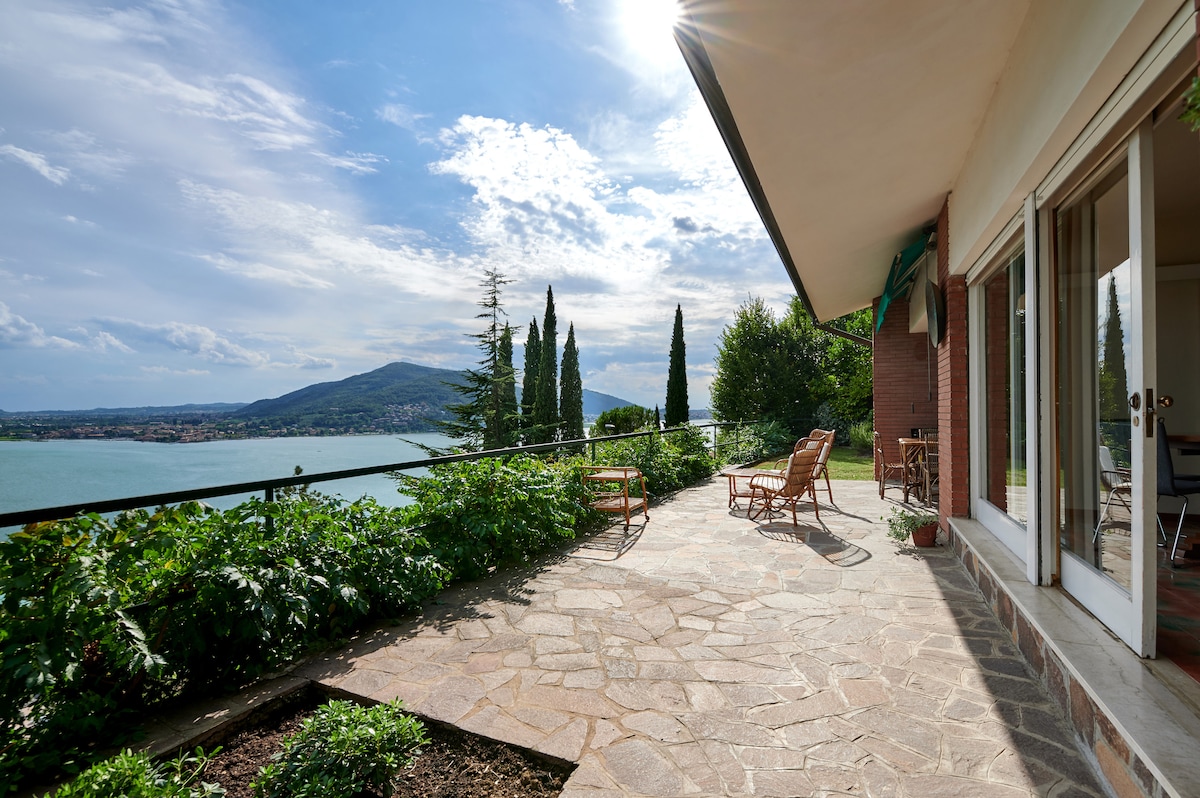An outdoor terrace features stone flooring and provides a panoramic view of the lake and surrounding hills. Comfortable lounge chairs are arranged for relaxation, with greenery providing a natural border. Large glass doors offer access to the interior space, allowing for an abundance of natural light.
