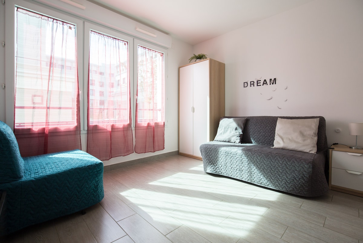 A bright living space with large windows dressed in sheer curtains allows natural light to fill the room. A turquoise convertible sofa and a gray convertible sofa provide seating options. A minimalist wardrobe and bedside table complete the simple decor, enhancing the open feel.