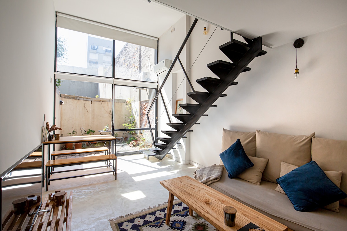A spacious living area features a light-filled atmosphere with large windows overlooking a private patio. A minimalist sofa with blue cushions faces a wooden coffee table. A stylish staircase with black metal railings leads to the upper floor, contributing to the open design.