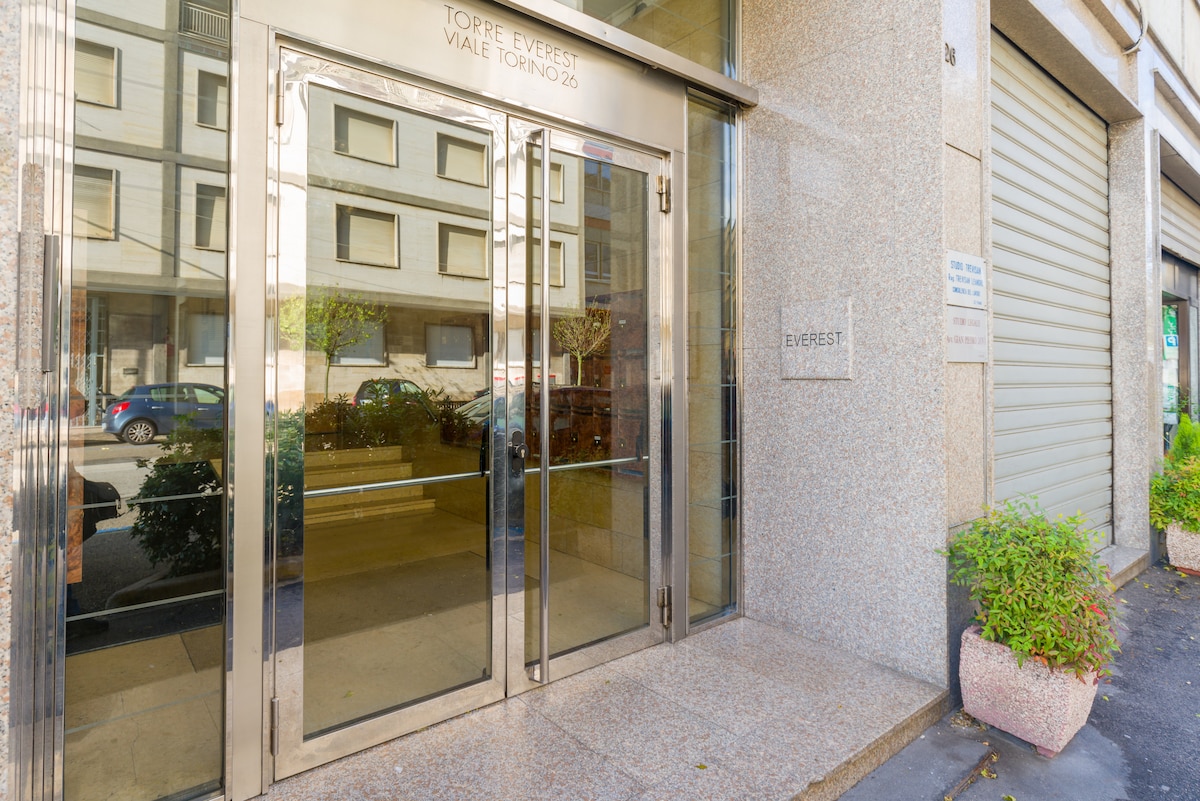 The entrance of the building is framed by large glass doors, reflecting the exterior surroundings. Marble finishes are prominent, enhanced by potted plants on either side of the entrance. Clear visibility of the address is displayed above the door.