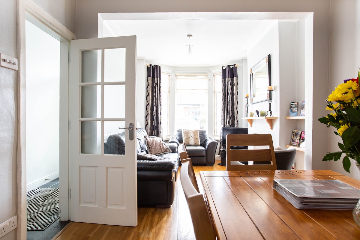 A bright living area is visible through open doors, featuring comfortable seating with cushions. Large windows allow natural light, while dark curtains add contrast. The dining space includes a wooden table with chairs, and a vase of flowers adds a touch of color on the table.