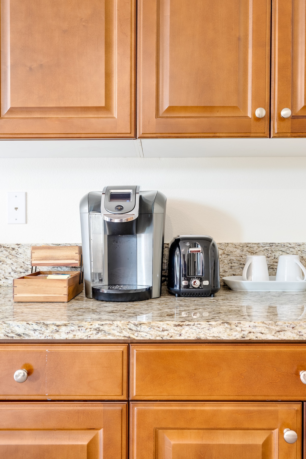 A kitchen countertop displays a coffee maker and toaster, both in modern designs. A wooden caddy holds assorted coffee supplies, while two white cups sit on a dish rack. The cabinetry features a warm wood finish, complemented by a granite surface.