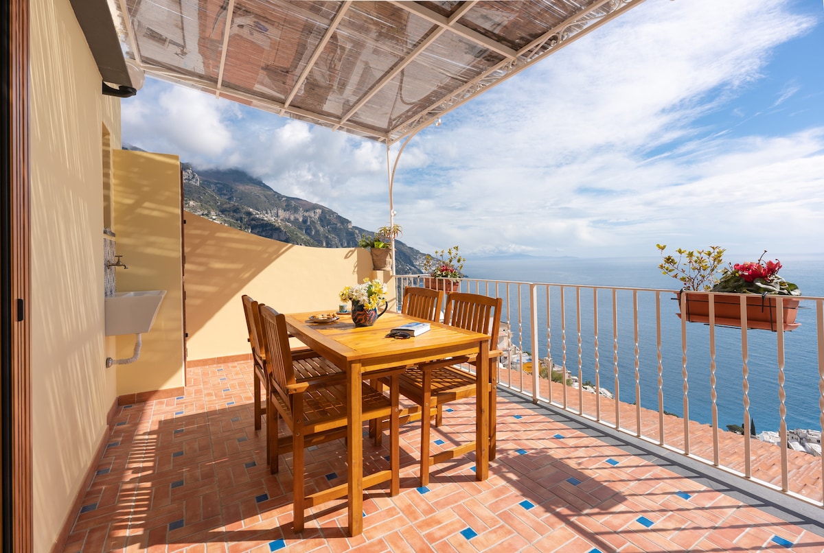 An outdoor terrace features a wooden dining table surrounded by matching chairs, offering panoramic views of the Amalfi Coast. Flower pots add a touch of greenery to the space. A pergola provides partial shade, and the scenic horizon blends sky and water.