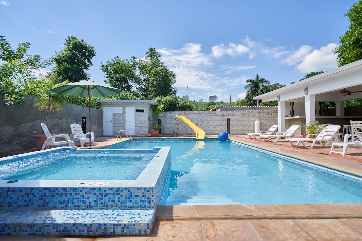 A refreshing pool area features a turquoise swimming pool with a water slide, surrounded by sun loungers and a shaded umbrella. A jacuzzi is positioned next to the pool, complemented by lush greenery and a clear blue sky above.