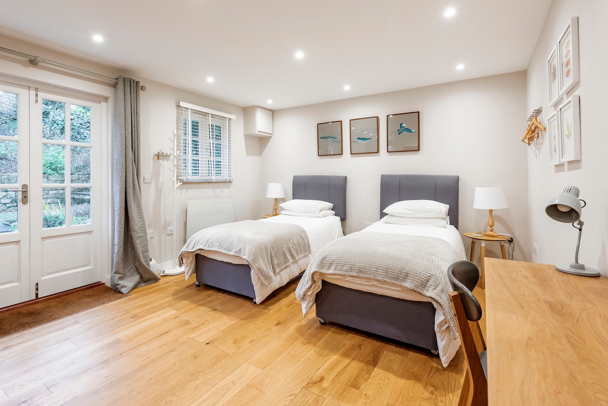 The spacious sleeping area features two single beds dressed in high-quality linens, with soft light illuminating the warm wooden floor. A well-lit desk is positioned beside one bed, while glass doors open to a tranquil outdoor view.