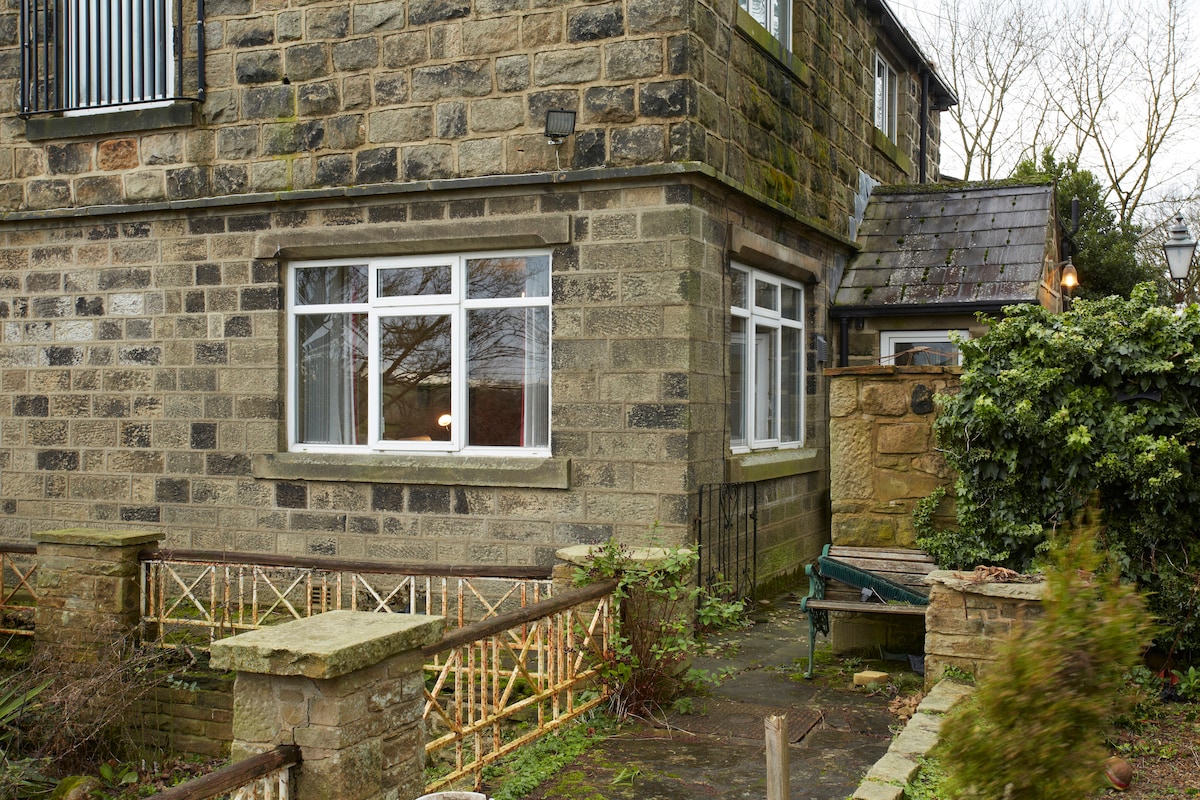 The exterior of a stone building is depicted, featuring a large, multi-paned window with a simple railing in front. The surrounding area includes a pathway and garden space, with greenery and a small bench visible at the side.