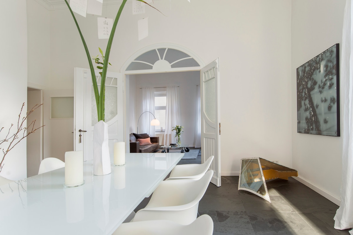 A spacious dining area is highlighted by a white table surrounded by sleek chairs. Natural light fills the room through large windows, illuminating a contemporary art piece on the wall and a cozy seating area visible beyond an archway.