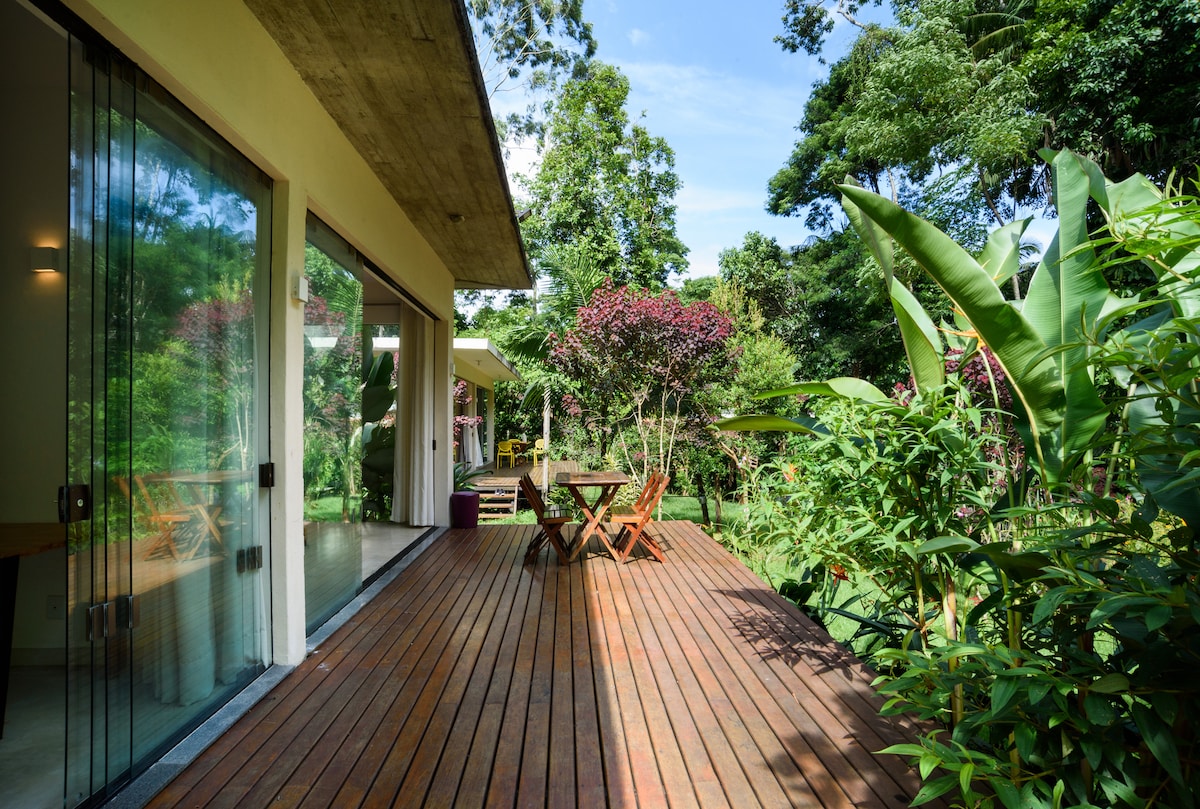 A spacious wooden deck extends from large glass doors, offering an unobstructed view of a vibrant garden filled with lush plants and flowering trees. Natural light floods the area, highlighting comfortable outdoor seating arrangements that invite relaxation amidst the surrounding greenery.