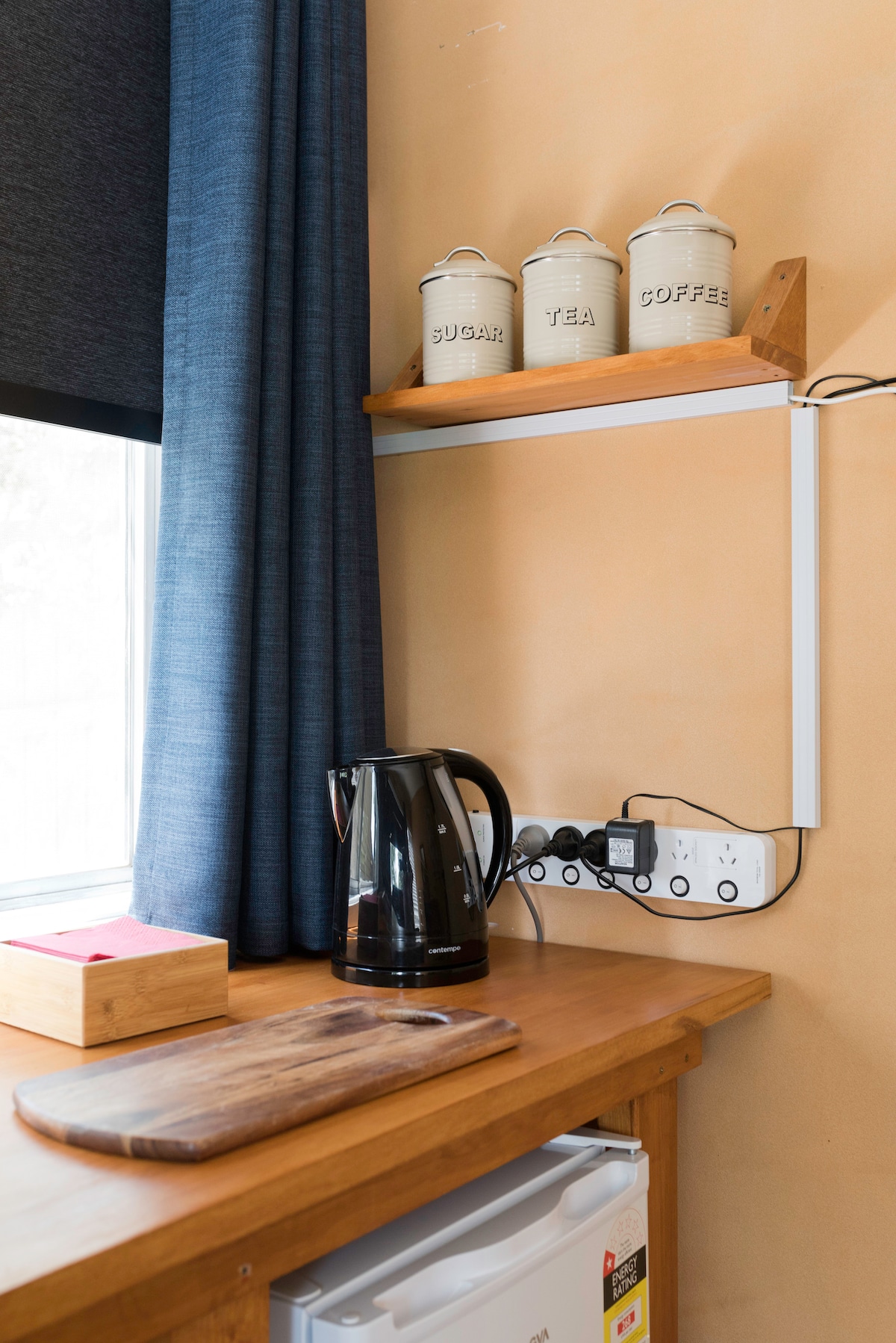A wooden countertop features a kettle, cutting board, and a storage box. A small white shelf holds three canisters labeled 'SUGAR,' 'TEA,' and 'COFFEE.' A window with dark curtains allows natural light to fill the space, enhancing the practical area.