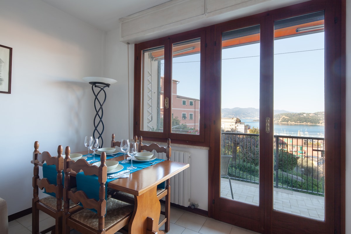 A dining area is visible, featuring a wooden table set for four with turquoise placemats and glassware. Large windows provide a view of the sea and surrounding scenery, allowing natural light to fill the space. A floor lamp adds illumination beside the table.