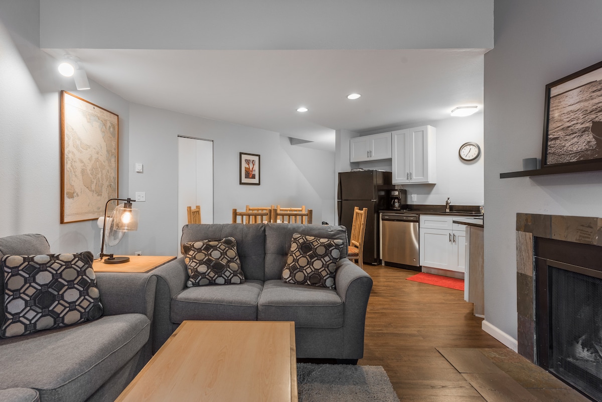 A modern living area features a comfortable gray sofa with patterned cushions. A wooden coffee table sits in front, and a dining area with wooden chairs is visible in the background. The kitchen, equipped with updated appliances, is located to the side of the space.