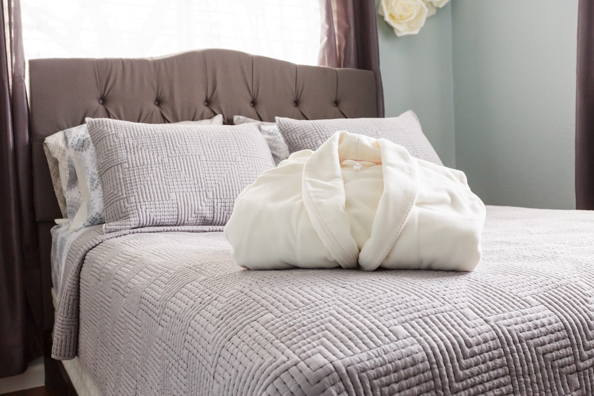A spacious bed with a quilted cover is featured, adorned with multiple pillows in coordinated patterns. A plush white robe is neatly placed in the center, inviting relaxation. Natural light filters through the windows, complemented by elegant dark curtains.